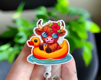 May include: A colorful badge reel featuring a cartoon Highland cow wearing sunglasses and a flower crown, floating in a yellow duck-shaped pool float. The cow is holding a drink with a straw. The badge reel has a white retractable cord and a clear plastic clip.
