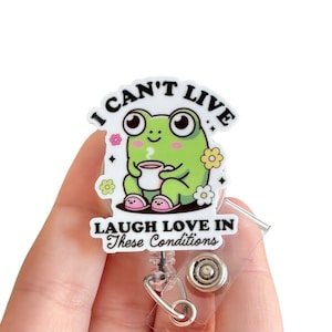 May include: A white plastic badge holder with a green frog cartoon holding a cup of coffee. The text on the badge holder reads "I can't live laugh love in these conditions".