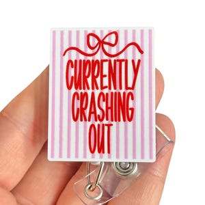 May include: A rectangular badge reel with a white and pink striped background and the words "CURRENTLY CRASHING OUT" in red. A red bow is at the top. The badge reel has a clear plastic strap and a metal clip.