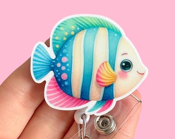 Könnte beinhalten: Ein farbenfroher Cartoon-Fisch mit blauen, weißen und rosa Streifen, grünen und gelben Akzenten und großen Augen. Der Fisch ist an einem durchsichtigen, einziehbaren Ausweishalter aus Kunststoff befestigt.