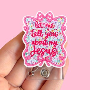 May include: A pink and white badge reel with the text "let me tell you about my Jesus." The badge has a floral design and a pink ribbon border. The reel has a clear plastic retractable cord and a metal clip.