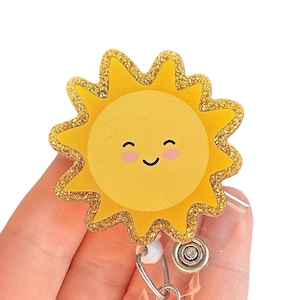 May include: A gold glitter sun with a smiling face and pink cheeks, attached to a clear plastic badge reel with a silver swivel clip.