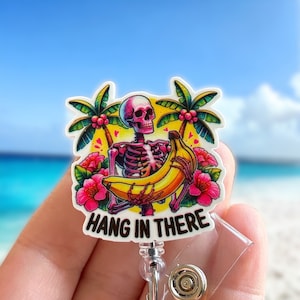 May include: A badge reel featuring a skeleton holding a banana, with palm trees and pink flowers against a yellow background. The text "HANG IN THERE" is displayed below the image. The reel has a clear plastic front and a metal clip.