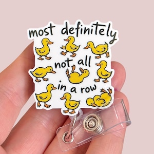 May include: A white badge reel with the text "most definitely not all in a row" and illustrations of yellow ducklings. The badge reel has a clear plastic strap and a metal clip.
