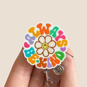 May include: A colorful badge holder with a white background and the words "Always Be Kind" in a rainbow of colors. A smiling yellow daisy with a white center is in the middle of the badge holder.