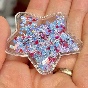 K&ouml;nnte beinhalten: Ein transparenter, sternf&ouml;rmiger Anh&auml;nger aus Kunststoff, gef&uuml;llt mit blauem, rotem und wei&szlig;em Glitzer. Der Glitzer hat die Form von Sternen und kleinen, d&uuml;nnen St&uuml;cken.