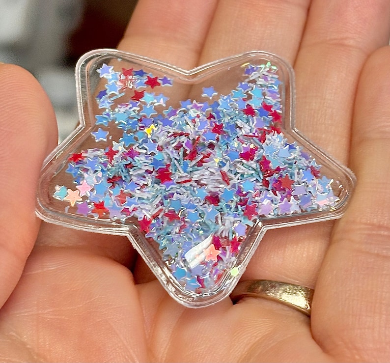 K&ouml;nnte beinhalten: Ein transparenter, sternf&ouml;rmiger Anh&auml;nger aus Kunststoff, gef&uuml;llt mit blauem, rotem und wei&szlig;em Glitzer. Der Glitzer hat die Form von Sternen und kleinen, d&uuml;nnen St&uuml;cken.