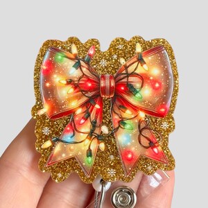 May include: A gold glitter badge reel with a red bow and colorful Christmas lights. The bow is decorated with a white snowflake pattern.
