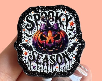 Op de afbeelding: Zwarte glitter badge reel met een spookachtig seizoensontwerp met een jack-o-lantern met een paarse strik, vleermuizen en sterren.