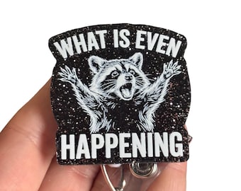 May include: A black glittery badge reel featuring a white illustration of a raccoon with its paws raised in the air. The text "WHAT IS EVEN HAPPENING" is printed in bold white capital letters above and below the raccoon graphic.