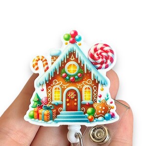 May include: A Christmas-themed badge reel featuring a gingerbread house design. The house is decorated with colorful candies, gifts, and a candy cane. The roof is covered in snow, and the house has a red door and yellow windows.