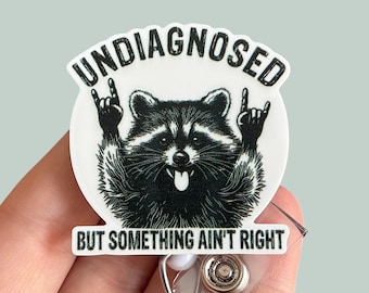 May include: A white and black badge reel featuring a raccoon illustration with the text "UNDIAGNOSED" above and "BUT SOMETHING AIN'T RIGHT" below. The raccoon is making a rock and roll hand gesture.