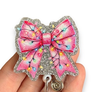 May include: A silver glitter badge reel with a pink bow and colorful lights. The bow is decorated with a sparkly, glittery finish.