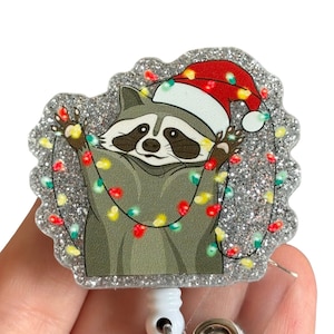 May include: A silver glitter badge reel with a cartoon raccoon wearing a Santa hat and tangled in Christmas lights.