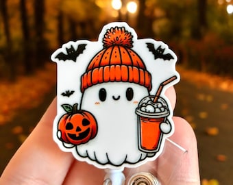 Puede incluir: Un fantasma blanco que lleva un gorro de punto naranja y rojo con un pompón blanco, sosteniendo una pequeña calabaza naranja y una taza roja y blanca con una pajita. El fantasma está en un portagajes retráctil de plástico transparente.