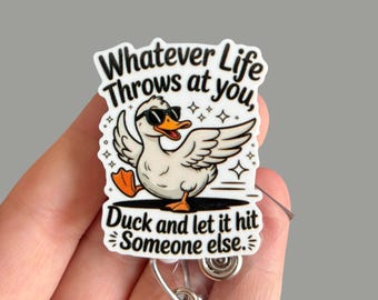 May include: A white badge with a cartoon duck wearing sunglasses and orange feet. The badge reads "Whatever Life Throws at you, Duck and let it hit Someone else." The badge is held by a person's hand.