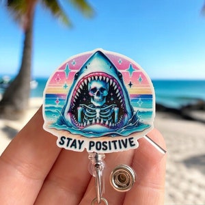 May include: A colorful badge reel with a graphic of a shark with a skeleton inside its mouth. The shark is in the ocean with a sunset in the background. The text "STAY POSITIVE" is printed below the shark.