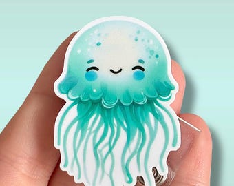 Könnte beinhalten: Eine türkisblaue und weiße Cartoon-Qualle mit einem lächelnden Gesicht. Die Qualle befindet sich auf einem durchsichtigen Plastik-Badgehalter mit einem Metallclip.