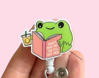 May include: A green frog cartoon character reading a pink book with the text "Don't bug me or I'll eat you". The frog is holding a cup of coffee with a smiley face. The cartoon is attached to a retractable badge holder.