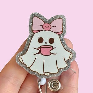 May include: A silver glitter ghost with a pink bow and a skull charm, holding a pink coffee cup. The ghost is attached to a clear plastic retractable badge holder.