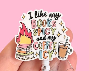 Può includere: Un portabadge bianco con una grafica colorata che dice "Mi piacciono i miei libri piccanti e il mio caffè ghiacciato". La grafica presenta una pila di libri con fiamme che escono dalla parte superiore e una tazza di caffè con una cannuccia.