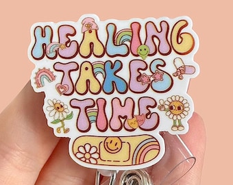 May include: A white and pastel-colored badge reel with the words "HEALING TAKES TIME" in a groovy font. The badge features rainbows, flowers, and smiley faces. The reel has a clear plastic retractable cord and a metal clip.
