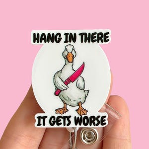 May include: White badge holder with a cartoon duck holding a pink knife. The text on the badge holder reads "Hang in there it gets worse".