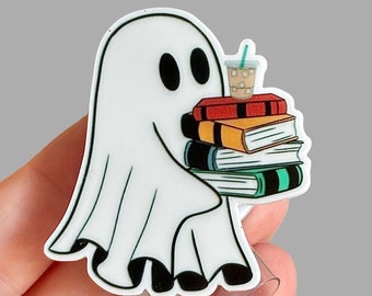May include: A white ghost-shaped badge reel with a stack of books and a drink. The ghost has black eyes and is holding the books and drink. The books are red, orange, and green. The drink has ice cubes and a green straw.
