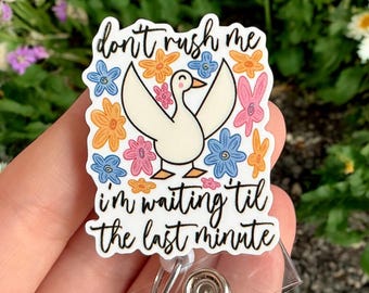 May include: A white badge reel with a cartoon duck and floral accents. The text reads "don't rush me, I'm waiting til the last minute." The badge reel has a clear plastic strap and a metal clip.