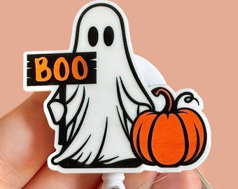 May include: A white ghost holding a sign that says "BOO" and a small orange pumpkin. The ghost and pumpkin are attached to a clear plastic retractable badge holder.