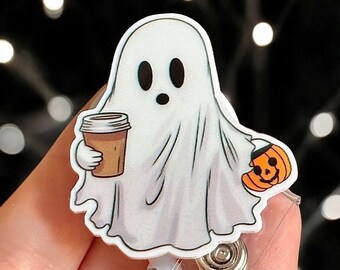 Op de afbeelding: Een witte, spookvormige badge reel met een cartoonontwerp. Het spook houdt een koffiekopje en een pompoen vast. De badge reel heeft een intrekbaar koord en een metalen clip.