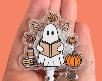 May include: A Halloween-themed badge reel featuring a white ghost reading a book, surrounded by orange fall leaves and a small orange pumpkin. A stack of books and a coffee cup are also included in the design.