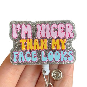 May include: Silver glitter badge holder with a white and blue retractable badge reel. The badge holder has the text "I'm nicer than my face looks" in colorful letters.