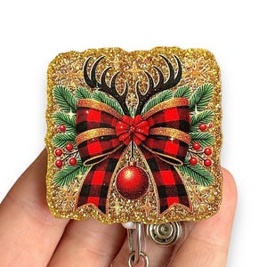 May include: Gold glitter square badge reel with a red and black plaid bow, gold antlers, and a red ornament. The badge reel has a clear plastic retractable cord.