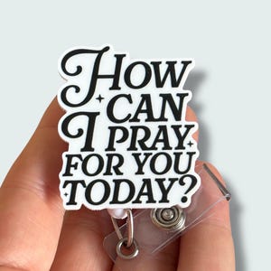 May include: A white and black badge reel with the text "How Can I Pray For You Today?" in a stylized font. The badge reel has a clear plastic holder and a metal clip.