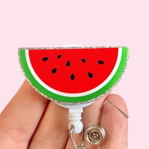 May include: A white retractable badge holder with a watermelon design. The watermelon is red with black seeds and a green rind. The holder has a silver glitter trim.