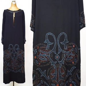 May include: A floor-length black dress featuring long sleeves and a keyhole neckline. The garment is decorated with intricate beadwork in shades of blue and rust, forming symmetrical patterns along the hemline and the cuffs of the sleeves.