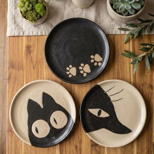 Puede incluir: Tres platos redondos de cerámica con diseños pintados a mano. Un plato negro muestra tres huellas de patas beige. Dos platos beige presentan caras de gato negras estilizadas, una con ojos grandes y redondos y otra de perfil.