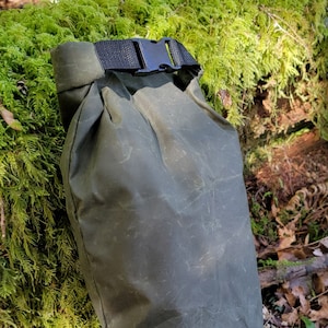 May include: A green waxed canvas dry bag with a black buckle closure. The bag is sitting on a bed of moss and leaves. The bag has a small blue label with the text "PNW Bushcraft" on it.