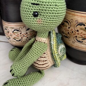 May include: A green crocheted turtle toy with brown and beige stripes on its shell. The turtle has black eyes and a stitched-on mouth.