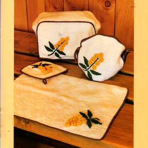 May include: A set of four crocheted pot holders with a wattle design. The pot holders are white with brown trim and feature yellow and green wattle flowers. The pot holders are arranged on a wooden surface. The text "Wattle" is printed in orange at the top of the image. The text "Lucia Russo" is handwritten in black at the bottom of the image.