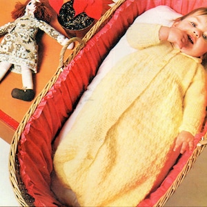 Könnte beinhalten: Ein gelbes gestricktes Babykleid mit Knopfleiste, getragen von einem Baby, das in einem Weidenkorb mit roter Auskleidung liegt.