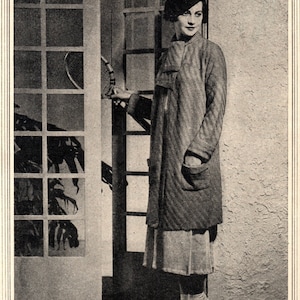 May include: A woman in a striped coat and a hat stands in a doorway holding a tennis racket. She is wearing a white skirt and white shoes.