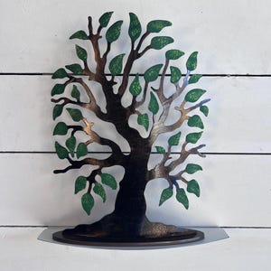 May include: A metal tree sculpture with a brown trunk and green leaves. The tree is standing on a brown base.