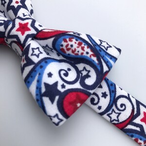 May include: A white bow tie with a paisley pattern in red, blue, and navy blue. The pattern also includes white stars.