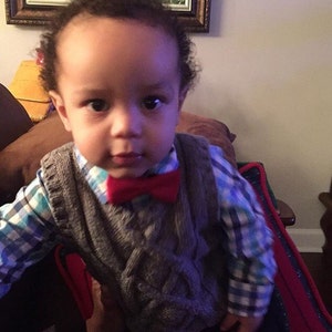 May include: A young boy wearing a gray cable knit vest with a red bow tie over a blue and white plaid shirt.