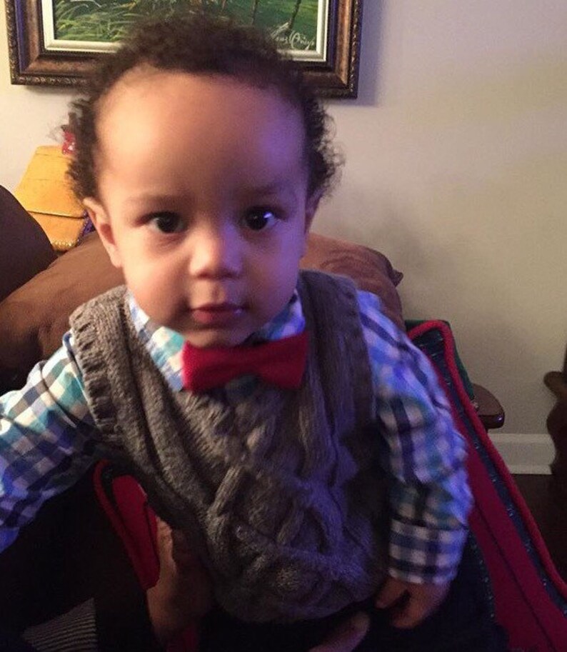 May include: A young boy wearing a gray cable knit vest with a red bow tie over a blue and white plaid shirt.