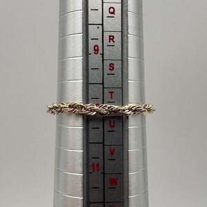 May include: A gold-toned rope-style ring is positioned on a silver-colored metal ring mandrel. The ring rests between the size markers Q and R on the measuring tool, which features clear black text and horizontal lines for sizing.