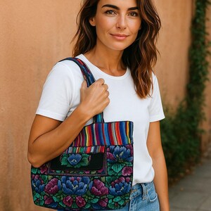 May include: A patterned tote bag featuring a floral design in blue, purple, and green. The bag has a black pocket and striped handles. The person is wearing a white t-shirt and blue jeans.