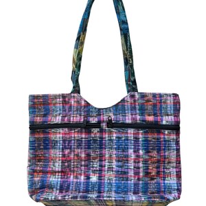 May include: A multi-coloured tote bag with a plaid design in blue, purple, pink, and green. The bag features two black zipped pockets and a matching patterned handle. The bag is designed for carrying items.
